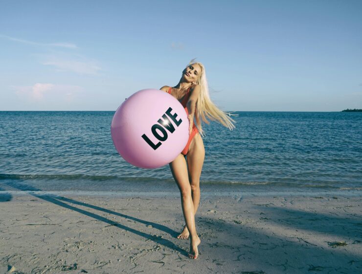 A woman holding a pink love-themed beach ball by the ocean, expressing freedom, softness and emotional openness.