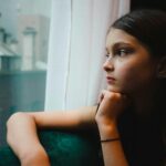 A thoughtful young woman gazing out of a window, reflecting quietly in a soft moment of emotional healing and inner awareness.