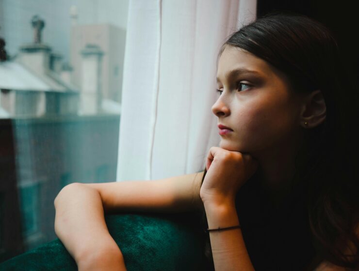 A thoughtful young woman gazing out of a window, reflecting quietly in a soft moment of emotional healing and inner awareness.