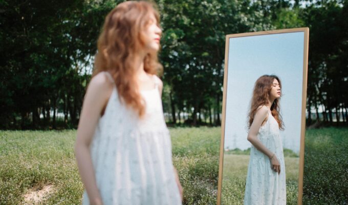 Young woman in a white dress standing by a mirror in a quiet field, reflecting on her life like the main character, SheJournal lifestyle image
