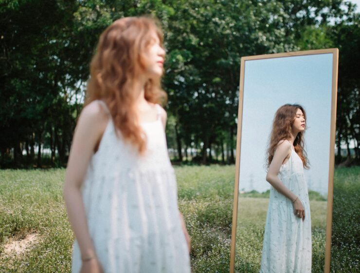 Young woman in a white dress standing by a mirror in a quiet field, reflecting on her life like the main character, SheJournal lifestyle image
